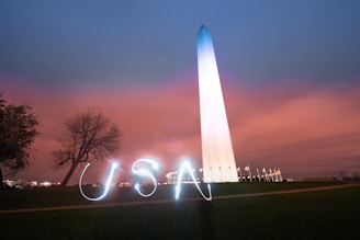 A tall, illuminated monument reaches into a sky painted with a gradient of blue and pink hues. In the foreground, the word 'USA' is creatively displayed using light painting. A leafless tree stands to the left, while a row of flags flutters to the right at the monument's base.