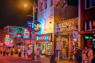 A lively street scene at night, featuring brightly lit neon signs and bustling crowds in front of B.B. King's Company Store. The buildings are adorned with colorful signage and the atmosphere is vibrant with a sense of nightlife excitement.