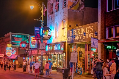 A lively street scene at night, featuring brightly lit neon signs and bustling crowds in front of B.B. King's Company Store. The buildings are adorned with colorful signage and the atmosphere is vibrant with a sense of nightlife excitement.