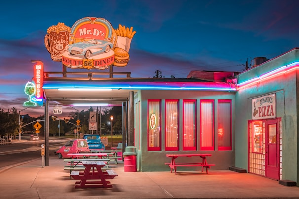 A vintage-style diner named Mr. D'z, located along Route 66, is depicted with retro neon signs and bold colors. The setting is during dusk, with a vibrant sky in the background. A classic car is parked outside, and there are red picnic tables and a bright sign for Armando's Pizza.
