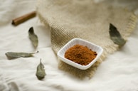 A small white square dish containing a heap of brown, finely ground spice is placed on a piece of textured burlap cloth. Surrounding the dish, there are several dried bay leaves and a cinnamon stick scattered across a cream-colored fabric surface.