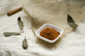 A small white square dish containing a heap of brown, finely ground spice is placed on a piece of textured burlap cloth. Surrounding the dish, there are several dried bay leaves and a cinnamon stick scattered across a cream-colored fabric surface.