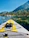 A paddleboard with bright yellow and gray traction pads is floating on a clear, calm lake. The board features a pair of black sandals secured by bungee cords at the front. In the background, a picturesque landscape displays dense, green pine trees lining the shore beneath a clear blue sky. Rocky formations are scattered along the shoreline, with mountains rising in the distance.