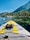 A paddleboard with bright yellow and gray traction pads is floating on a clear, calm lake. The board features a pair of black sandals secured by bungee cords at the front. In the background, a picturesque landscape displays dense, green pine trees lining the shore beneath a clear blue sky. Rocky formations are scattered along the shoreline, with mountains rising in the distance.