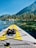 A paddleboard with bright yellow and gray traction pads is floating on a clear, calm lake. The board features a pair of black sandals secured by bungee cords at the front. In the background, a picturesque landscape displays dense, green pine trees lining the shore beneath a clear blue sky. Rocky formations are scattered along the shoreline, with mountains rising in the distance.