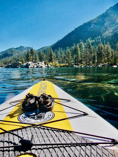 A paddleboard with bright yellow and gray traction pads is floating on a clear, calm lake. The board features a pair of black sandals secured by bungee cords at the front. In the background, a picturesque landscape displays dense, green pine trees lining the shore beneath a clear blue sky. Rocky formations are scattered along the shoreline, with mountains rising in the distance.