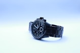 A sleek black wristwatch with a metal band is presented against a light gradient background. The watch face is detailed with several dials and hands, marked with numbers and indices. The brand name is visible on the watch face.