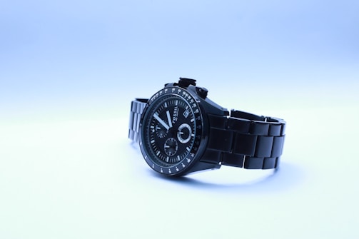 A sleek black wristwatch with a metal band is presented against a light gradient background. The watch face is detailed with several dials and hands, marked with numbers and indices. The brand name is visible on the watch face.