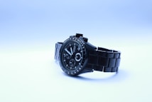 A sleek black wristwatch with a metal band is presented against a light gradient background. The watch face is detailed with several dials and hands, marked with numbers and indices. The brand name is visible on the watch face.