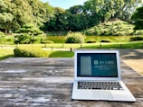 A laptop displaying Japanese text is placed on a weathered wooden table in an outdoor setting. Behind the table, there is a scenic park with neatly trimmed bushes, a small pond, and lush green trees. The sky is clear, indicating a sunny day.