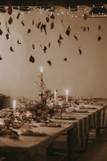 A rustic dining table elegantly set with plates, glasses, and decorative foliage. Candles cast a warm glow, while dried leaves hang from above as part of the ceiling decor. Twinkling lights add a whimsical touch, enhancing the cozy atmosphere.