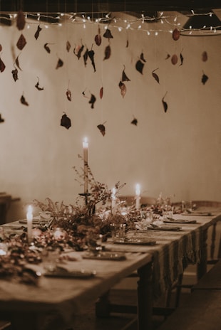 A rustic dining table elegantly set with plates, glasses, and decorative foliage. Candles cast a warm glow, while dried leaves hang from above as part of the ceiling decor. Twinkling lights add a whimsical touch, enhancing the cozy atmosphere.