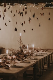 A rustic dining table elegantly set with plates, glasses, and decorative foliage. Candles cast a warm glow, while dried leaves hang from above as part of the ceiling decor. Twinkling lights add a whimsical touch, enhancing the cozy atmosphere.