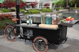 A black tricycle cart is set up in an outdoor space with various colorful bottles and trays of fresh produce displayed on its countertop. The cart features a modern design with tan leather saddle and wheels, and the surroundings show neatly arranged greenery and red-leafed trees. An urban setting forms the backdrop with people in the distance.