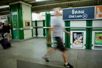 A slightly blurred person walks past a sign at the Chit Lom station, with visible advertisements on the walls. The station features green and white interior details, and another person is visible with luggage in the background.