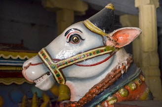 A detailed statue of a white bull adorned with colorful decorations including a vibrant harness and painted ornaments. The bull has distinct large eyes, curved horns, and an intricately designed collar. It is placed in an indoor setting with muted lighting.