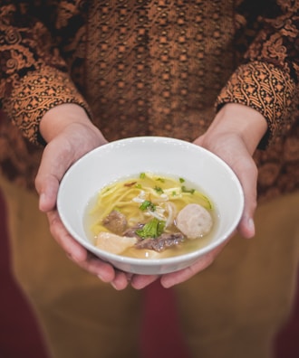 A hand pouring savory broth into a bowl filled with bubur ayam and toppings.