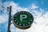 A circular green sign with a large letter 'P' in the center, encircled by the words 'Municipal Parking'. The sign is mounted on a metal pole with attached wiring, set against a backdrop of a bright blue sky with scattered white clouds.