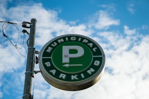 A circular green sign with a large letter 'P' in the center, encircled by the words 'Municipal Parking'. The sign is mounted on a metal pole with attached wiring, set against a backdrop of a bright blue sky with scattered white clouds.