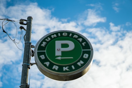 A circular green sign with a large letter 'P' in the center, encircled by the words 'Municipal Parking'. The sign is mounted on a metal pole with attached wiring, set against a backdrop of a bright blue sky with scattered white clouds.