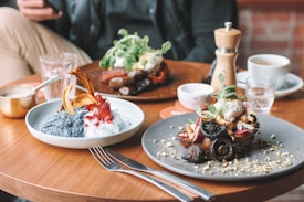 A table is set with plates of gourmet food, including a dish with mushrooms, greens, and possibly poached eggs on one plate, and a dessert with chia seeds, pomegranate seeds, sliced fruit, and cream on another. There are also various utensils, glasses, a pepper grinder, coffee cups, and a hand holding a phone in the background.
