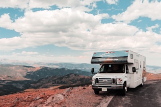 A large recreational vehicle is parked on the side of a mountain road, offering a panoramic view of expansive mountain ranges under a partly cloudy blue sky. The terrain is rocky with some patches of grass, creating a rugged landscape around the vehicle.