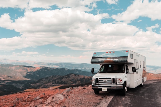 A large recreational vehicle is parked on the side of a mountain road, offering a panoramic view of expansive mountain ranges under a partly cloudy blue sky. The terrain is rocky with some patches of grass, creating a rugged landscape around the vehicle.
