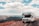 A large recreational vehicle is parked on the side of a mountain road, offering a panoramic view of expansive mountain ranges under a partly cloudy blue sky. The terrain is rocky with some patches of grass, creating a rugged landscape around the vehicle.