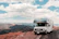 A happy RV traveler checking mail on a laptop beside their parked RV at a scenic mountain campground.