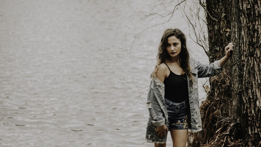 woman in black spaghetti strap top and blue denim jacket standing near tree with body of water behind