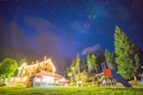 A brightly lit house at night surrounded by trees, with a clear starry sky above. The building has a warm, welcoming glow, and there is a playground with a slide nearby on a well-kept lawn.