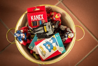 A yellow metal container holds several colorful superhero action figures, a box of Kanex colored pencils, and what appears to be a puzzle or card game. The figures are arranged haphazardly, and the overall atmosphere suggests a playful and nostalgic theme.