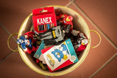 A yellow metal container holds several colorful superhero action figures, a box of Kanex colored pencils, and what appears to be a puzzle or card game. The figures are arranged haphazardly, and the overall atmosphere suggests a playful and nostalgic theme.