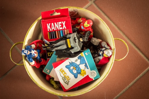 A yellow metal container holds several colorful superhero action figures, a box of Kanex colored pencils, and what appears to be a puzzle or card game. The figures are arranged haphazardly, and the overall atmosphere suggests a playful and nostalgic theme.