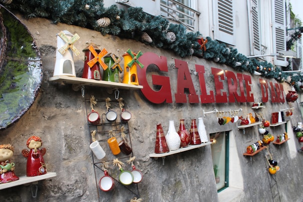 Various decorative ceramic objects are displayed on shelves attached to a stone wall, including colorful vases, windmill models, and hanging cups. The wall features the bold red lettering 'GALERIE D'ART' and is adorned with a garland of pine branches and pinecones, creating a festive atmosphere. Shutters and windows suggest a building exterior.