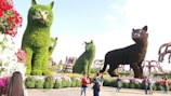 Several large, cat-shaped topiaries are displayed in a vibrant garden setting, crafted meticulously from green foliage. The cats vary in size and exhibit intricate details such as distinct feline faces. Surrounding these topiary sculptures are various brightly colored flowers including red, pink, and white blooms. People are seen walking and observing the display, capturing photos and enjoying the scenic landscape.