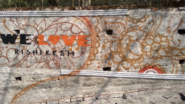 A wall mural features intricate floral and paisley designs with earthy colors like orange and brown. In the foreground, the phrase 'We Love Rishikesh' is painted, with 'Love' styled as a heart symbol. The background is a textured gray wall.