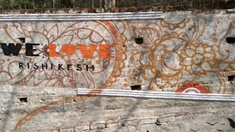 A wall mural features intricate floral and paisley designs with earthy colors like orange and brown. In the foreground, the phrase 'We Love Rishikesh' is painted, with 'Love' styled as a heart symbol. The background is a textured gray wall.