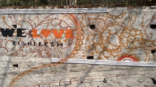 A wall mural features intricate floral and paisley designs with earthy colors like orange and brown. In the foreground, the phrase 'We Love Rishikesh' is painted, with 'Love' styled as a heart symbol. The background is a textured gray wall.