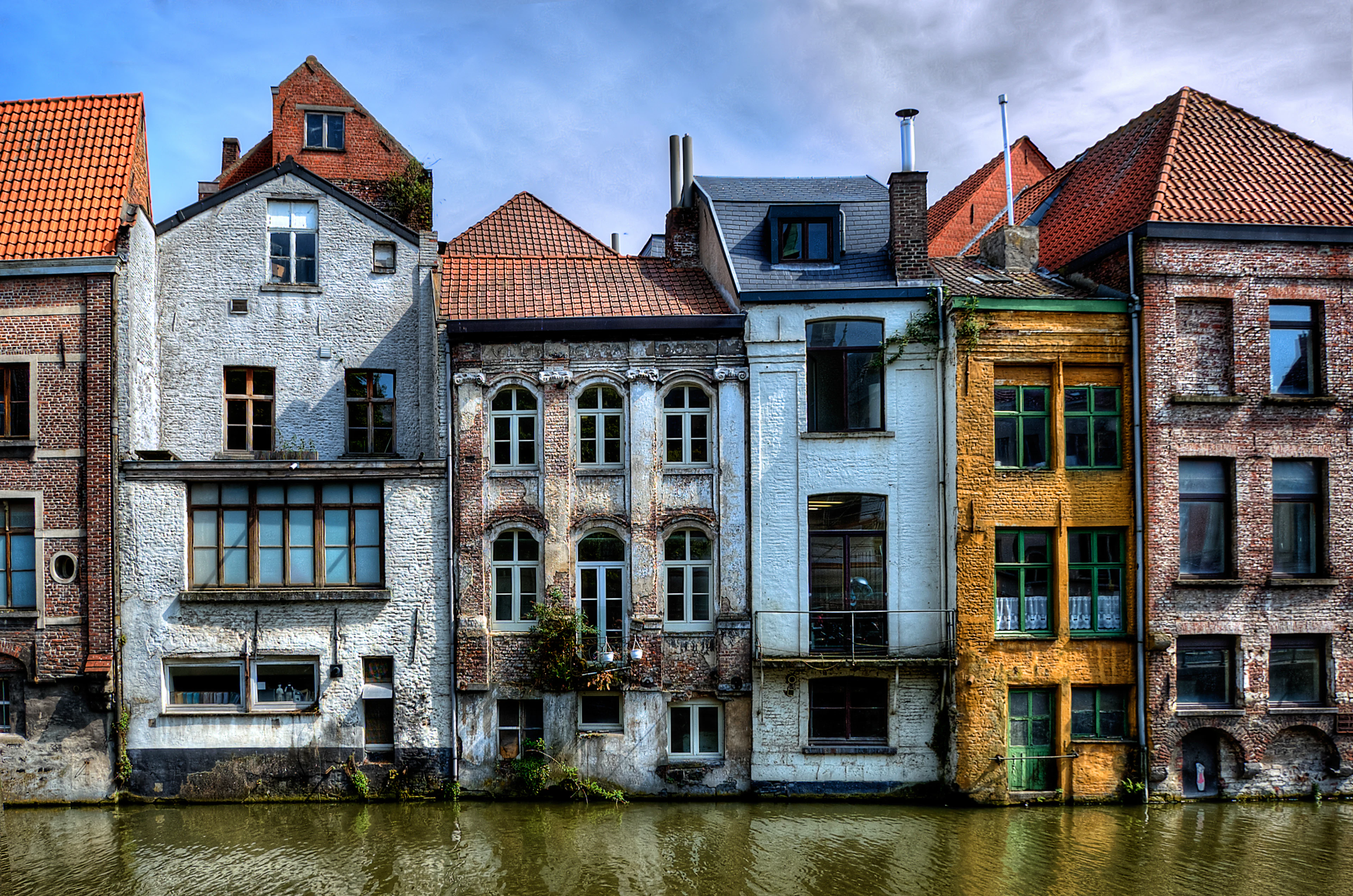 Weathered brick and plaster rear facades in white, orange and blue rising straight out of the canal water, seen from the Kraanlei in central Ghent