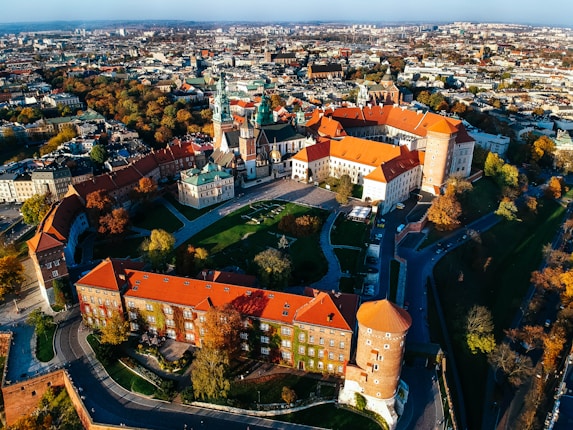 An aerial view of a historical castle complex with red-tiled roofs, surrounded by lush greenery and trees with autumn foliage. The architecture is a blend of medieval and Renaissance styles, featuring towers and a spacious courtyard.