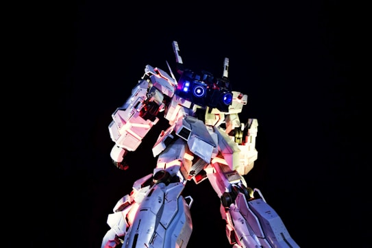 A towering, highly detailed robot stands against a dark background. It is illuminated by bright lights, with glowing blue and pink accents highlighting its mechanical features. The robot appears imposing and sophisticated in design, with a focus on angular panels and intricate components.