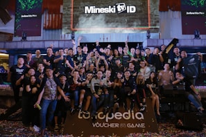 A large group of people is gathered together in a celebratory atmosphere, posing for a group photo. Many are making peace signs or giving thumbs up. Above the group, there is a large screen with the text 'Mineski TV' displayed. The area is decorated with colorful confetti scattered on the ground.