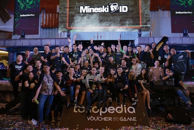 A large group of people is gathered together in a celebratory atmosphere, posing for a group photo. Many are making peace signs or giving thumbs up. Above the group, there is a large screen with the text 'Mineski TV' displayed. The area is decorated with colorful confetti scattered on the ground.