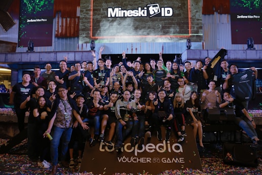 A large group of people is gathered together in a celebratory atmosphere, posing for a group photo. Many are making peace signs or giving thumbs up. Above the group, there is a large screen with the text 'Mineski TV' displayed. The area is decorated with colorful confetti scattered on the ground.