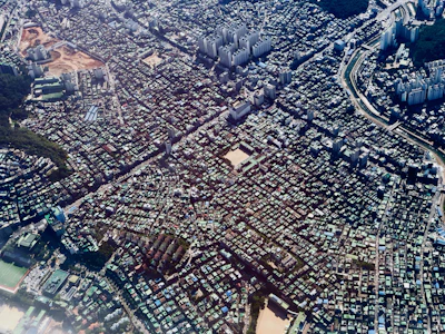 An aerial view of an urban landscape with a dense arrangement of buildings and structures. The city features a mix of high-rise apartments and smaller residential buildings, intersected by roads and possibly a river. Vegetation is visible in sections, hinting at parks or wooded areas.