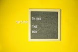 A grey felt letter board with a wooden frame is placed against a solid yellow background. The board displays the words 'THINK THE BOX' in white letters, while the word 'OUTSIDE' is placed on the yellow background to the left of the board in similar white letters.