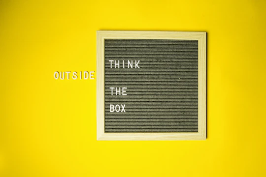 A grey felt letter board with a wooden frame is placed against a solid yellow background. The board displays the words 'THINK THE BOX' in white letters, while the word 'OUTSIDE' is placed on the yellow background to the left of the board in similar white letters.