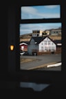 A view through a window reveals a small collection of buildings, including a quaint hotel with a traditional architectural style and red and white accents. The buildings are situated on a paved road with a barren landscape in the background, under a partly cloudy sky.