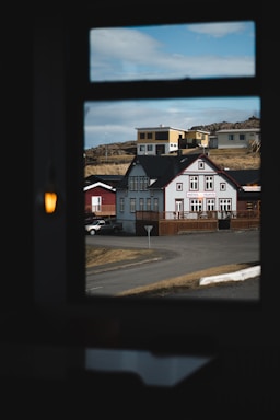 A view through a window reveals a small collection of buildings, including a quaint hotel with a traditional architectural style and red and white accents. The buildings are situated on a paved road with a barren landscape in the background, under a partly cloudy sky.
